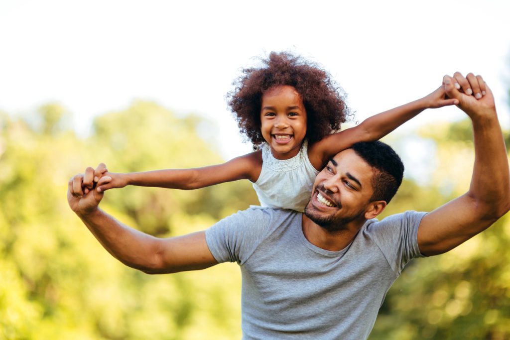 A man carries his daughter on his back on a sunny day, their hands interlocked and their arms outstretched.