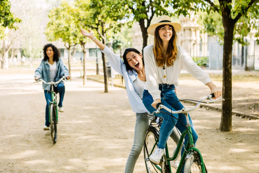 Three women laughing and riding bikes in a park during summer.