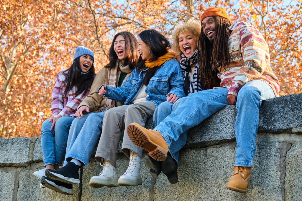 A group of friends hanging out and laughing. They are dressed in layers and sitting on a wall in front of some autumn trees.